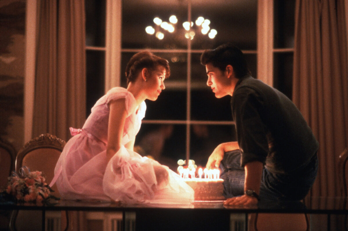 Two teens sitting on a table romantically looking at each other over a lit birthday cake.
